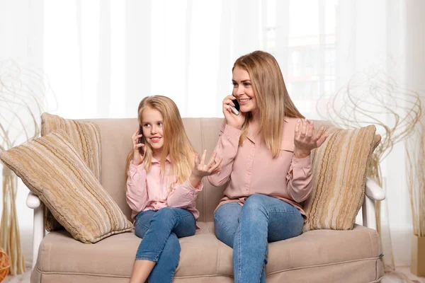 Shot of a mom and daughter of blondes with long hair wearing pink shirts and jeans sitting on the couch at home and having fun talking on the phone. Daughter imitates mom\'s behavior