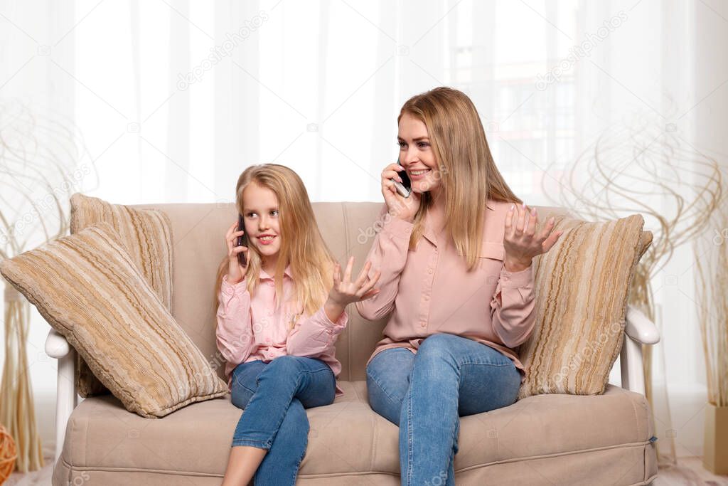 Shot of a mom and daughter of blondes with long hair wearing pink shirts and jeans sitting on the couch at home and having fun talking on the phone. Daughter imitates mom's behavior