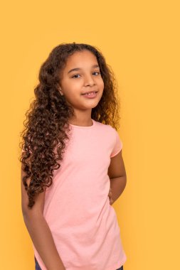 Koyu tenli, uzun kıvırcık saçlı, sarı arka planda pembe gömlekli genç bir kızın belden yukarı portresi.