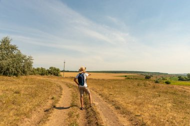 Hasır şapkalı bir sırt çantalı genç bir çocuğun kırsalda yolda yürürken ve manzaranın güzelliğine hayran kalırken görüntüsü.. 