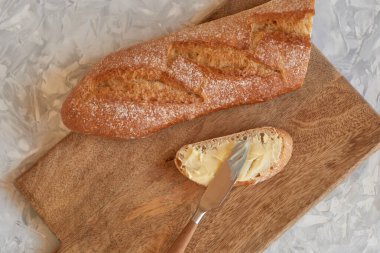 Gri masadaki ahşap bir tahtanın üzerinde, tereyağlı bir bıçakla bir dilim beyaz ekmeğin yakın çekimi, üst manzara. Yakınlarda bir baget var..