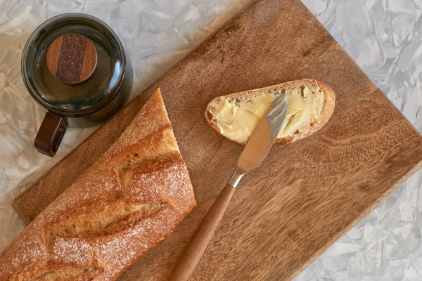 Gri masadaki ahşap bir tahtanın üzerinde, tereyağlı bir bıçakla bir dilim beyaz ekmeğin yakın çekimi, üst manzara. Yakınlarda baget ve çayın yarısı uzanır.