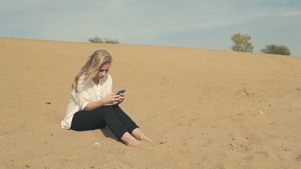 Босоногая девушка сидит на песке с телефоном — стоковое видео