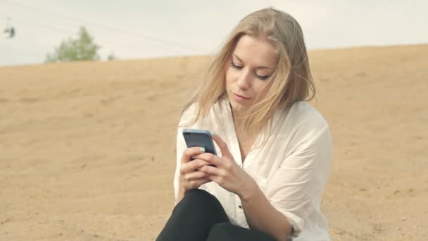 Gadis yang duduk di pasir dengan latar belakang gerbong kabel dengan telepon — Stok Video