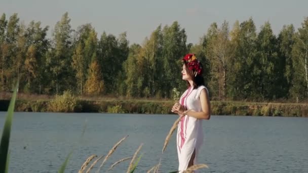 Schönes Mädchen in russischer Nationaltracht mit einem Kranz auf dem Kopf, der auf der Brücke am See steht — Stockvideo