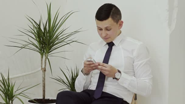 Empresario en camisa blanca y corbata, sentado en una silla y viendo las noticias en su teléfono — Vídeo de stock