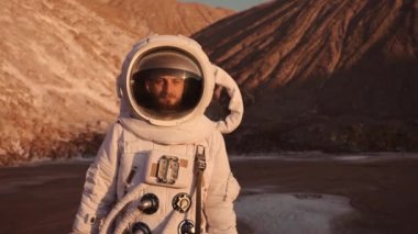 Astronot tepelerin yakınında dikkatli bir şekilde yürüyor ve önüne bakıyor. Güneş parlıyor.