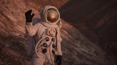 Astronot bir kaya üzerinde durup el sallıyor ve baş parmağını kaldırıyor. Güneş parlıyor.