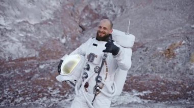 Elinde miğfer olan astronot geniş bir şekilde gülümsüyor ve başparmağını kaldırıyor.