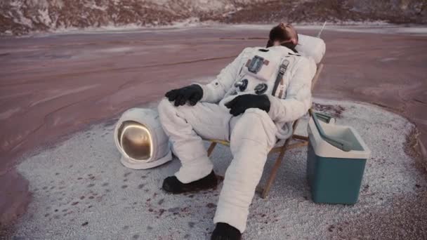 Buzdolabı olan kasksız bir astronot tepelerde bir sandalyede dinleniyor. — Stok video