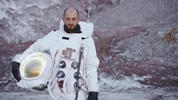 Astronauta se para con un casco en la mano junto a las colinas y mira a la cámara — Vídeo de stock