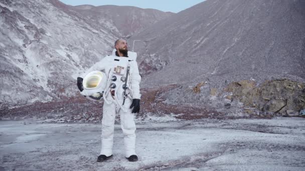 Astronaute se tient debout avec un casque à la main près des collines et regarde sur le côté — Video