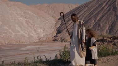 İsa elini su deposuna doğru tutan kızla yürür. Güneş parlıyor