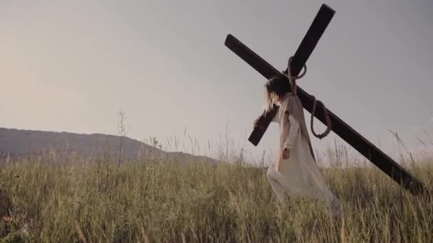 Иисус несет крест на спине. прогулки по траве, на фоне холмов. — стоковое видео