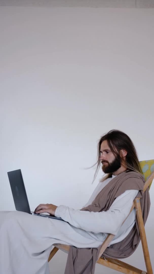 Jesús se sienta en un schizlong y trabaja en una computadora sobre un fondo blanco. Estudio. — Vídeo de stock