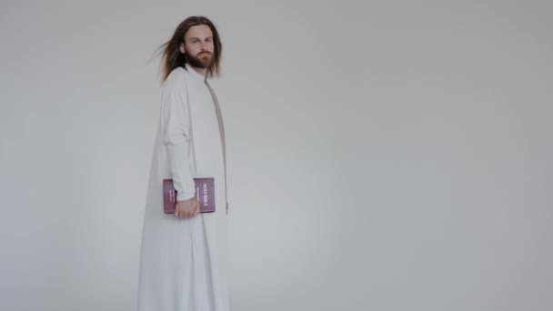 Иисус смотрит в камеру, держа Библию в руках на белом фоне — стоковое видео