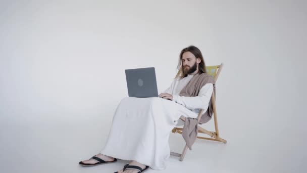Ο Ιησούς κάθεται σε ένα σχιζοφρενή και εργάζεται σε έναν υπολογιστή σε ένα λευκό φόντο. Στούντιο. — Αρχείο Βίντεο