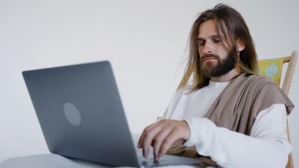 Jezus zit in een schizofreen en werkt op een computer op een witte achtergrond. Studio. — Stockvideo