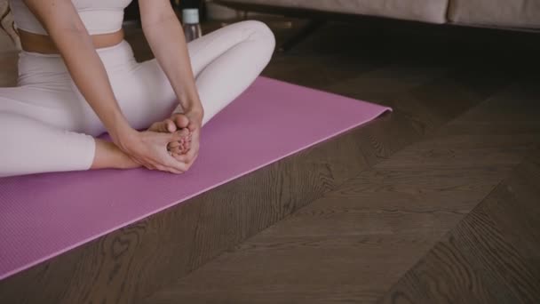 Fiatal szőke ül a jóga szőnyegen, összehajtott lábakkal a kezével. — Stock videók