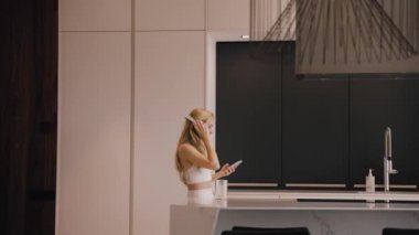 Mutfakta elinde telefon ve kulaklıkla dans eden genç bir kadın.