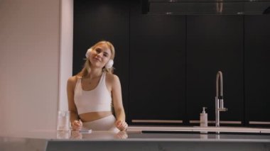 Kulaklıklı genç bir sarışın mutfakta dans edip mutlu bir şekilde müzik dinliyor.