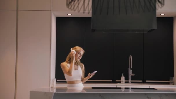 Uma jovem dançando com um telefone em suas mãos e fones de ouvido na cozinha — Vídeo de Stock