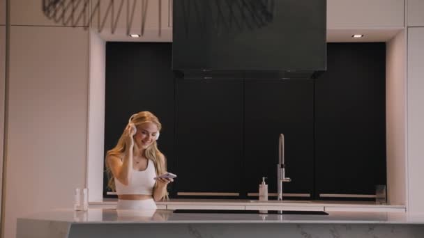 Una joven bailando con un teléfono en las manos y auriculares en la cocina — Vídeos de Stock