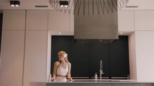 Een jonge vrolijke blonde vrouw dansend met hoofdtelefoon in de prachtige keuken — Stockvideo
