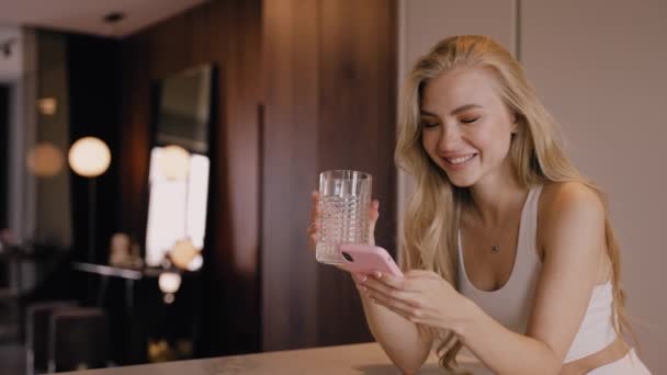 En ung blondin i köket dricker vatten ur ett glas och chattar per telefon — Stockvideo