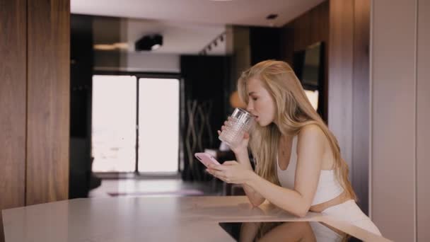 Una joven rubia en la cocina sonriendo, bebe agua y charla por teléfono — Vídeos de Stock