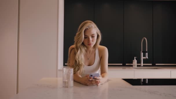 Een jonge blonde vrouw met een telefoon in haar handen kletst en drinkt water — Stockvideo
