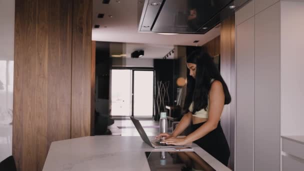 En ung mörkhyad brunett i det vackra köket skriver på en laptop — Stockvideo