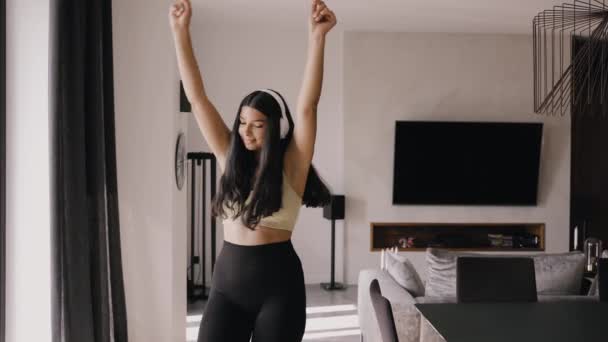 Молодая брюнетка в наушниках танцует в гостиной — стоковое видео