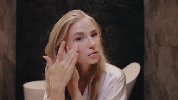 Ung blondin i en vit morgonrock i badrummet gäller grädde i ansiktet, hudvård — Stockvideo