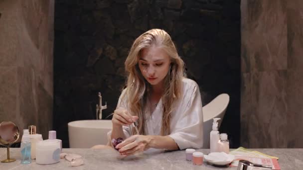 Young blonde in a white robe in the bathroom applies cream to her face, skincare — Stock Video