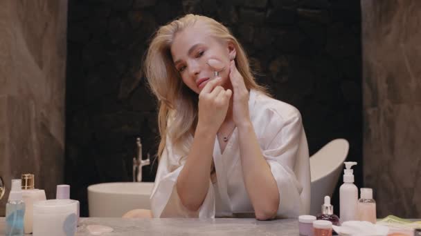 Молодая блондинка в белом халате в ванной комнате с роллер-массажером — стоковое видео