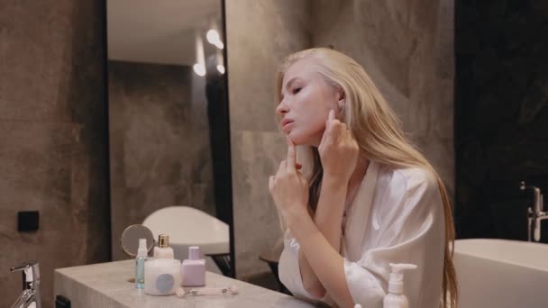 Молодая блондинка в белом халате в ванной комнате с массажным камнем — стоковое видео