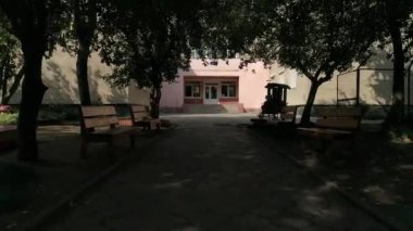 Ağaçları olan boş bir anaokulu binası. Ağaçların dalları girişe giden kaldırımın üzerinde asılı.