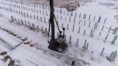 Yığın delici makinenin hava aracı görüntüsü güçlendirilmiş beton yığını kabloyla kaldırıyor. İşçi, yığının doğru yere yerleştirilmesine yardım eder. Kış bulutlu gün
