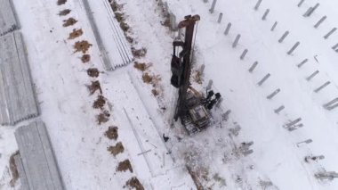 Kışın inşaat alanında çalışan bir kazık delici makinenin havadan drone görüntüsü. Yüksek çarpışma ağırlık sürücüleri yerin derinliklerine yığılıyor. Bölgede pekçok güçlendirilmiş beton yığını var. Kış bulutlu gün