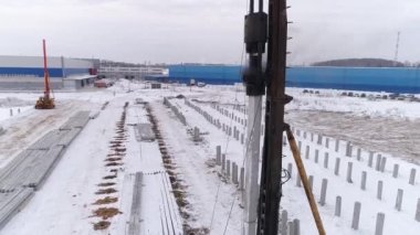 Kış şantiyesinde çalışan bir kazık delici makinenin hava aracı görüntüsünü kapatın. Yüksek çarpışma ağırlık sürücüleri yerin derinliklerine yığılıyor. Bölgede pekçok güçlendirilmiş beton yığını var. Kış bulutlu gün