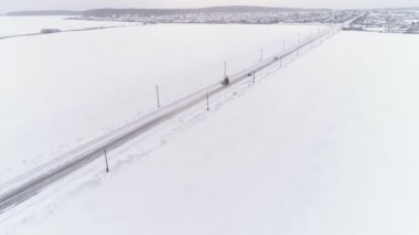 Sarı traktörün, karla kaplı kır yolunda tarlaların arasından geçtiği hava manzarası. Yolun kenarlarında elektrik direkleri ve kablolar var. Arabalar yol boyunca sürüyorlar. Kış bulutlu gün