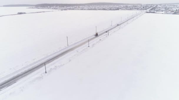 雪上のバケツに乗って黄色のトラクターの空中ビューフィールドを介してカントリーロードをカバー 道路の両側に電柱やワイヤ 車が道路に沿って運転している 冬曇りの日 — ストック動画