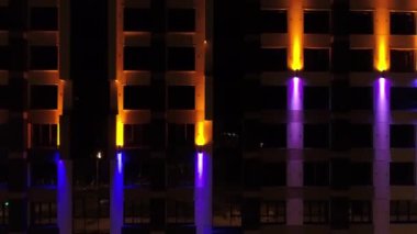 Güzel renkli mimari aydınlatması olan modern bir bina. Renkler değişir. İyi geceler, hava aracı görüntüsü. Kamera bina boyunca yükseliyor.