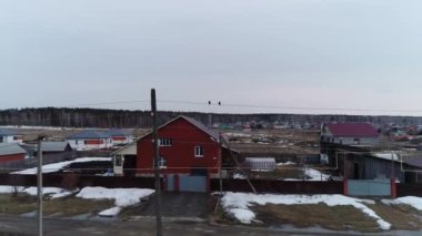 İki kuş (sığırcıklar) köyde kabloların üzerinde otururlar. Bahar bulutlu bir gün. Kar henüz erimedi ki. Arka planda sebze bahçeleri olan özel evler. Kamera kuşlara doğru uçuyor. Drone görünümü.