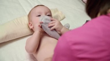 Masöz bebeğe karın masajı yapar (oğlan). Bebek sırtüstü yatar ve elinde yumuşak bir oyuncak tutar..