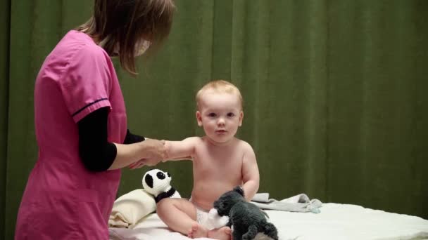 女按摩师给一个快乐的孩子 孩子的肩膀和手被按摩了一下 婴儿坐在那里 手里拿着一个柔软的玩具 — 图库视频影像