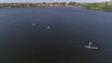 Stand-up paddle surf yapan insanların hava görüntüsü ve taşra kentindeki gölette ayakta yüzen sörfçüler (SUP). Kıyı 'da kilise, eski fabrika ve ağaçlar var. Yaz güneşli bir gün