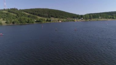 Stand-up paddle sörfü yapan insanların hava görüntüsü ve kalkış sörfü (SUP) ve taşra kentindeki gölette kayaklarda. Kilise, eski fabrika, ağaçlar ve sahilde şehir plajı var. Yaz güneşli bir gün