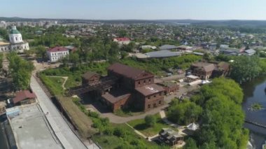 Şehrin kenar mahallelerinde nehir ve göletin yanındaki ağaçların arasında demir çatılı eski kırmızı tuğla fabrikasının hava manzarası. Yakınlarda eski evler ve kilise var. Yaz güneşli bir gün